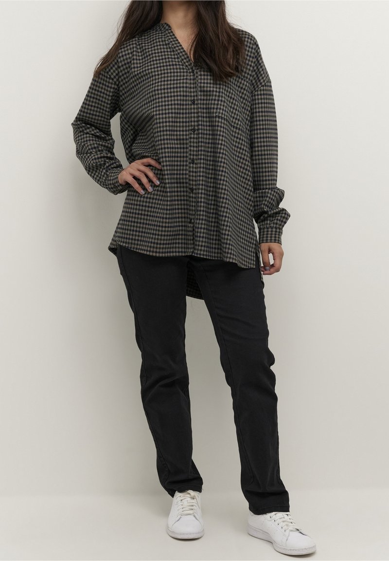 Femme portant une chemise à manches longues à carreaux gris et noirs, un pantalon noir et des baskets blanches, debout avec une main sur la hanche.