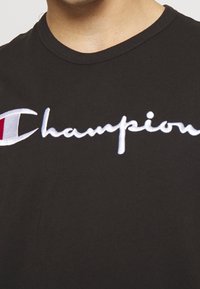 Mustat t-paita pyöreällä kaula-aukolla, jossa on valkoinen brodeerattu "Champion"-logo ja pieni punainen yksityiskohta logon vasemmalla puolella.