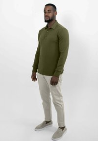 Man staat in een olijfgroene lange mouwen polo, beige broek en lichtgrijze instappers tegen een witte achtergrond.