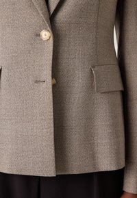 Hnědý strukturovaný blazer s dvěma velkými knoflíky, klopami a bočními kapsami. Přiléhavý střih s minimálními švy.