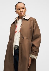 Νεαρή γυναίκα με πλεκτά μαλλιά που φοράει καφέ παλτό πάνω από λευκό πουλόβερ με ροζ και κόκκινα σχέδια και σκούρο τζιν με διακοσμητική αγκράφα ζώνης.