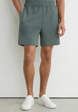Mand iført grønne teksturerede shorts med elastik i taljen og hvide sneakers, stående mod en hvid væg med afslappede hænder.
