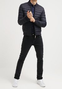 Man i svart puffjacka, mörk skjorta, svarta jeans och vita sneakers, som justerar sin vänstra handled med höger hand framför en enhetlig bakgrund.