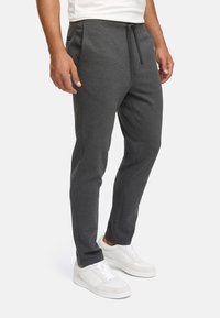 Man wearing dark gray slim-fit joggers with side pockets and white sneakers, standing against a white background.