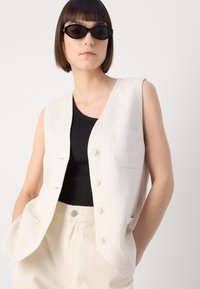 Mouwloos beige linnen gilet met vijf knopen, enkel zakje aan de linkerkant, gecombineerd met een zwarte aansluitende top en lichtgekleurde broeken.