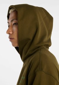 Felpa con cappuccio verde oliva dalla texture liscia, caratterizzata da un ampio cappuccio e dettagli di cuciture visibili. Design minimalista.