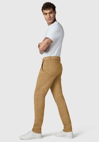 Homme tatoué portant un t-shirt blanc, un pantalon beige et des baskets blanches, debout les bras croisés, regardant sur le côté devant un fond uni.