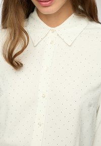 Blusa bianca con colletto, caratterizzata da un delicato motivo di piccoli punti argento. Chiusura frontale con bottoni, con una texture liscia e una vestibilità rilassata.