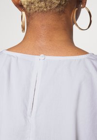 Top de tecido branco com decote arredondado, pequeno fecho com botão nas costas e detalhe de fenda vertical, apresentando uma aparência suave e texturizada.