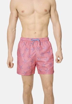 Hombre sin camiseta con pantalones cortos de baño rosa con estampados de estrellas de mar azules y cordón azul, de pie contra un fondo liso.