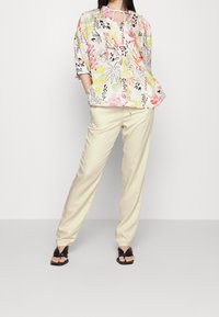 Bloemenprint shirt in pasteltinten met een relaxte pasvorm en striknek, gecombineerd met beige broeken en zwarte geknoopte sandalen.