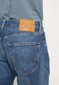 Jean en denim bleu avec une étiquette en cuir portant les détails "Levi Strauss & Co." et "Lot 502". Surpiqûres jaunes et poches arrière arrondies.