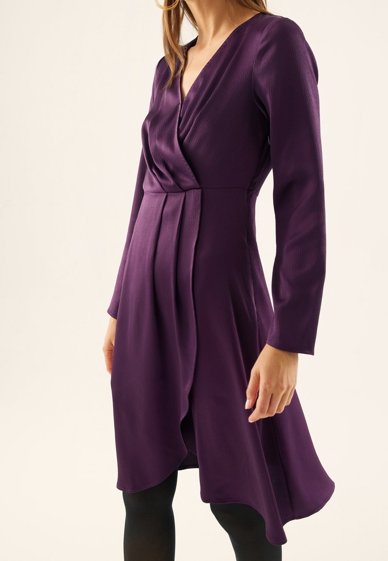 Femme portant une robe portefeuille violette à manches longues avec des plissés, associée à des collants noirs, sur un fond neutre uni.