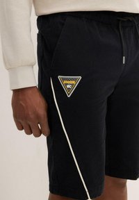 Pantaloni corti neri in tessuto, con una striscia laterale bianca e una toppa triangolare con "UNION GC" in giallo e nero. Vita elastica.