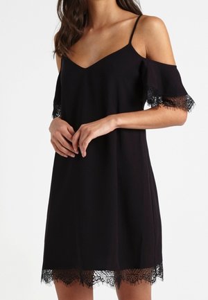 Kobieta w czarnej sukience typu cold-shoulder z koronkową lamówką na rękawach i dole, stojąca z rękami luźno złączonymi.