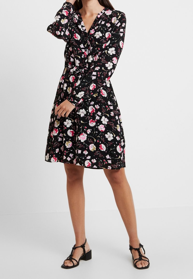 Femme portant une robe noire à fleurs à manches longues et des sandales noires à talons, debout devant un fond uni.