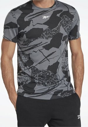 Mężczyzna w szaro-czarnej abstrakcyjnie wzorzystej koszulce Reebok i czarnych spodniach Reebok, ręce w kieszeniach, na jednolitym tle.