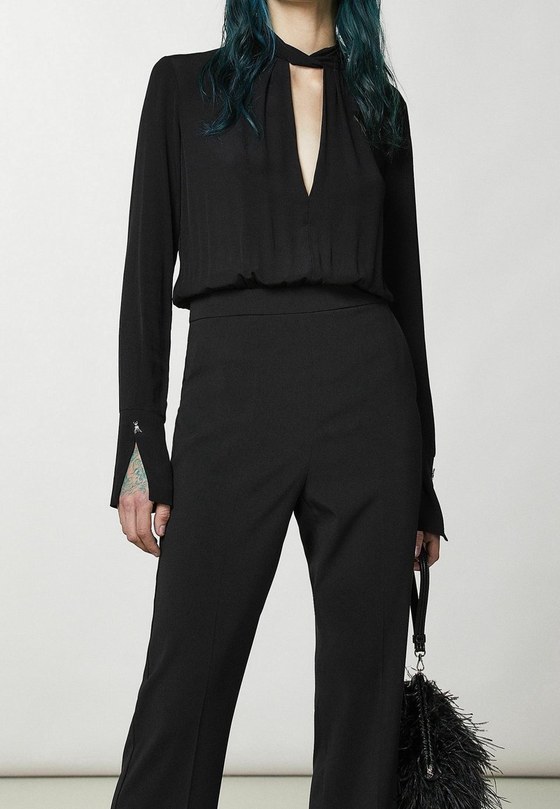 Femme aux cheveux bleu sarcelle portant un haut noir à manches longues avec une encolure en forme de trou et un pantalon noir taille haute, tenant un sac à main noir duveteux.