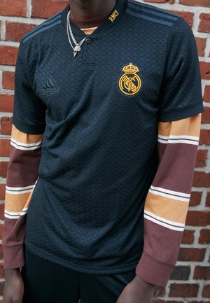 Personne portant un maillot de football noir du Real Madrid par-dessus une chemise à manches longues rayée marron et beige, debout contre un mur en briques rouges.