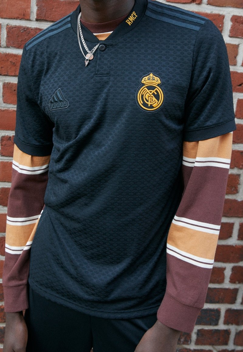 Personne portant un maillot de football noir du Real Madrid par-dessus une chemise à manches longues rayée marron et beige, debout contre un mur en briques rouges.