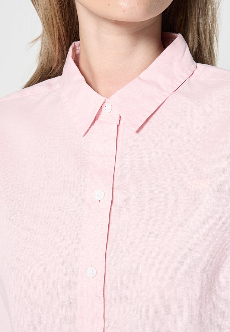 Camisa de botón de color rosa claro con cuello, hecha de un tejido suave. Presenta botones blancos y un pequeño logo de la marca en el pecho.