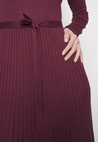 Robe côtelée bordeaux à manches longues, avec une taille nouée et une texture lisse, mettant en valeur des plis verticaux sur la jupe.