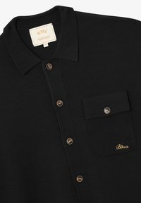 Zwarte langemouwen polo met een bruine knoop, borstzak met een knoop en geborduurd "Blkvis" logo in gouden draad.