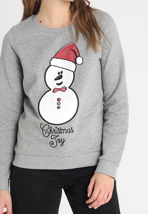 Kobieta w szarym swetrze z uśmiechniętym bałwankiem w czapce Mikołaja, czerwonym muszką i napisem „Christmas Joy” poniżej.