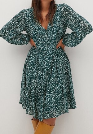 Kobieta ubrana w zieloną, długorękawową sukienkę w kwiaty z dekoltem w kształcie litery V, o długości do połowy uda, oraz beżowe kozaki do kolan, stojąca z rękami na biodrach.