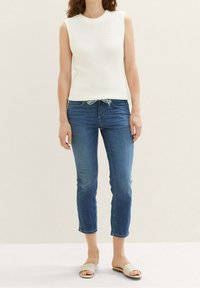 Mujer con top de punto blanco sin mangas, jeans cortos azules con cintura ajustable y sandalias beige, de pie contra un fondo liso.