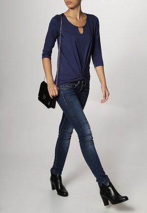 Navyblauwe, lange mouw top met een sleutelgatdetail en een knoop bij de taille, gecombineerd met donkere skinny jeans en zwarte enkellaarsjes met hak.