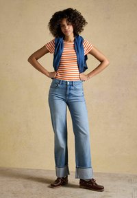 Jeune femme aux cheveux bouclés, debout les mains sur les hanches, portant une chemise rayée orange et blanche, un jean bleu avec des revers, des chaussures marron et un gilet bleu.