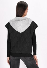 Gilet trapuntato nero con cappuccio grigio, caratterizzato da maniche corte e un motivo testurizzato. Il design è abbinato a un top nero a maniche lunghe sotto.