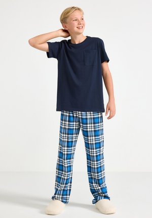 Marineblaues Baumwoll-T-Shirt mit Tasche, kombiniert mit blauen karierten Flanell-Schlafanzughosen. Model trägt beige flauschige Hausschuhe und steht in einem neutralen Raum.