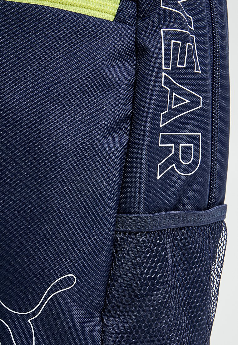 Mochila de tela azul marino con un zipper amarillo, logo de Puma blanco en la parte delantera, texto YEARN y un bolsillo lateral de malla.