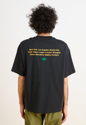 Personne aux cheveux bouclés portant un T-shirt noir avec la liste des grandes villes en texte jaune et un logo Adidas vert, pantalon vert olive.