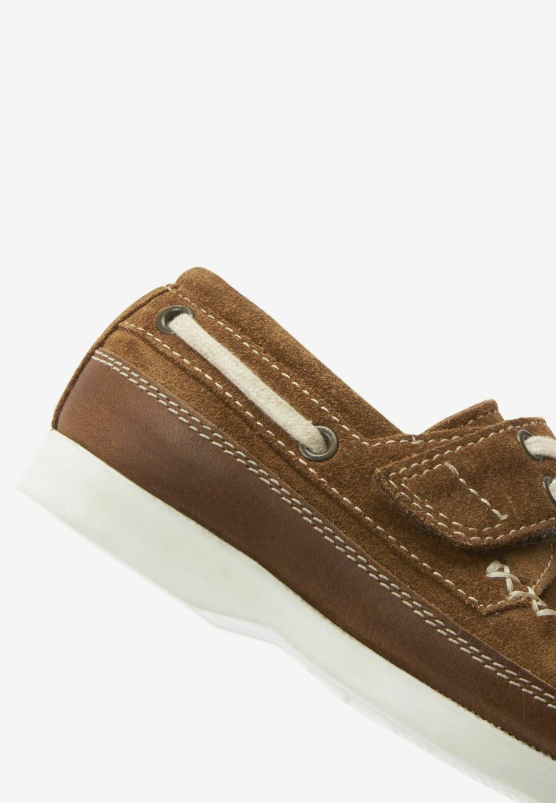 Chaussure en suède marron avec semelle en caoutchouc blanc. Présente des accents en cuir beige, des lacets beige et des coutures contrastées. Design à coupe basse.