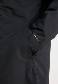 Manche de veste imperméable noire avec logo « DRYVENT », dotée d'un poignet zippé et d'une main dans la poche latérale.