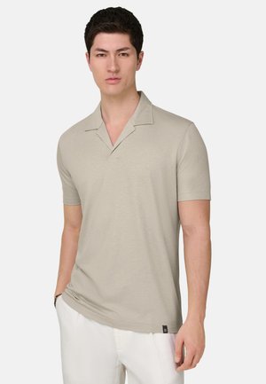 Jeune homme portant un polo beige à manches courtes avec un col cranté et un pantalon blanc, debout devant un fond uni.