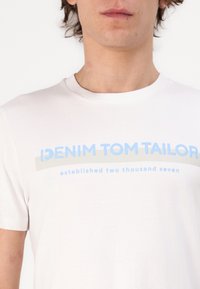 Valkoinen puuvillapaita, jossa on sininen ja harmaa graafinen tekstisuunnittelu. Teksti lukee "DENIM TOM TAILOR" ja "perustettu vuonna kaksituhatta seitsemän."