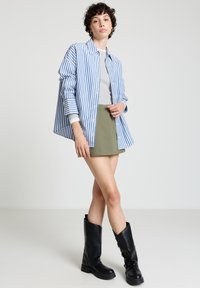 Blau-weiß gestreiftes Hemd über einem grauen Oberteil, kombiniert mit einem grünen Minirock und kniehohen schwarzen Stiefeln.