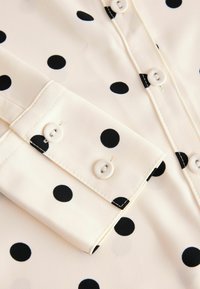 Blouse crème à pois noirs, avec un col et des poignets à boutons, ornés de deux boutons blancs. Tissu doux avec une texture lisse.