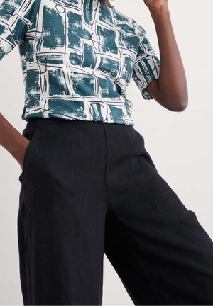 Vrouw die een teal- en witpatroon korte mouw top draagt, ingestopt in een zwarte, wijde, hoog-uitgesneden broek, poserend met één hand in de zak.