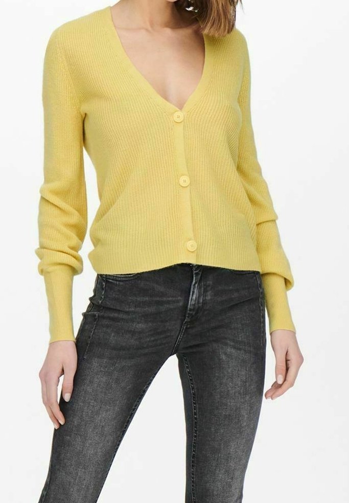 Žlutý žebrovaný pletený cardigan s výstřihem do V a pěti ladícími knoflíky, doplněný o těsné tmavé džíny.