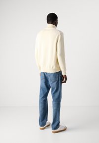 Cremefarbenes Strickpullover mit hohem Kragen, kombiniert mit hellblauen Jeans mit geradem Bein und weißen Sneakern mit hellbraunen Gummisohlen.