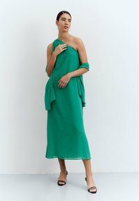 Vestido verde de un solo hombro en tejido ligero, con un cuerpo drapeado y falda fluida, que cuenta con una abertura lateral y un suave complemento de chal.