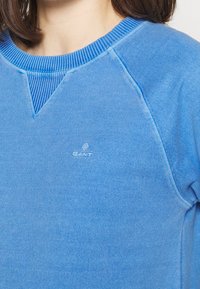 Sudadera azul con cuello acanalado, detalle en forma de V y pequeño logo de GANT en el pecho. Confeccionada en tejido suave y texturizado.