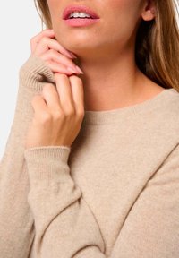 Pull en tricot beige avec une texture douce, doté d'un col rond et de longues manches. Des mains tiennent délicatement l'encolure, mettant en valeur le détail des manches.