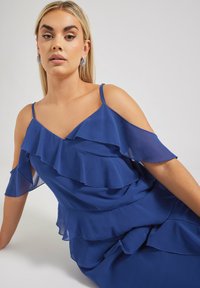 Blå lagerklänning med volangdetaljer, tunna axelband och off-shoulder design. Tillverkad av lättviktsmaterial med en mjuk textur.