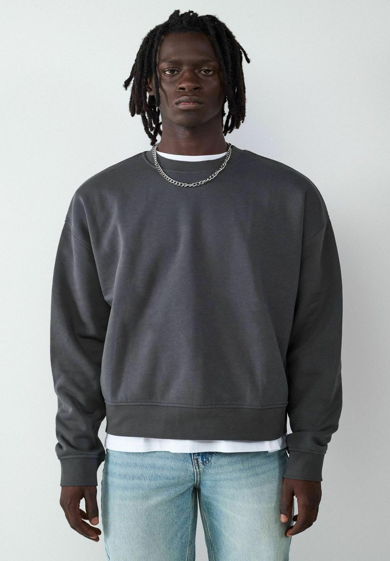 Ciemnoszary sweatshirt z długimi rękawami, okrągłym dekoltem, ściągaczami przy mankietach i u dołu. Nosi się go na białej koszuli, w zestawieniu z jasnoniebieskimi dżinsami.